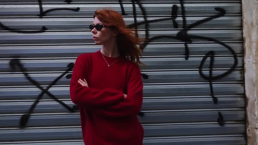 A stylish redhead woman in sunglasses and a red sweater poses confidently in front of a graffiti-covered metal door on an urban street