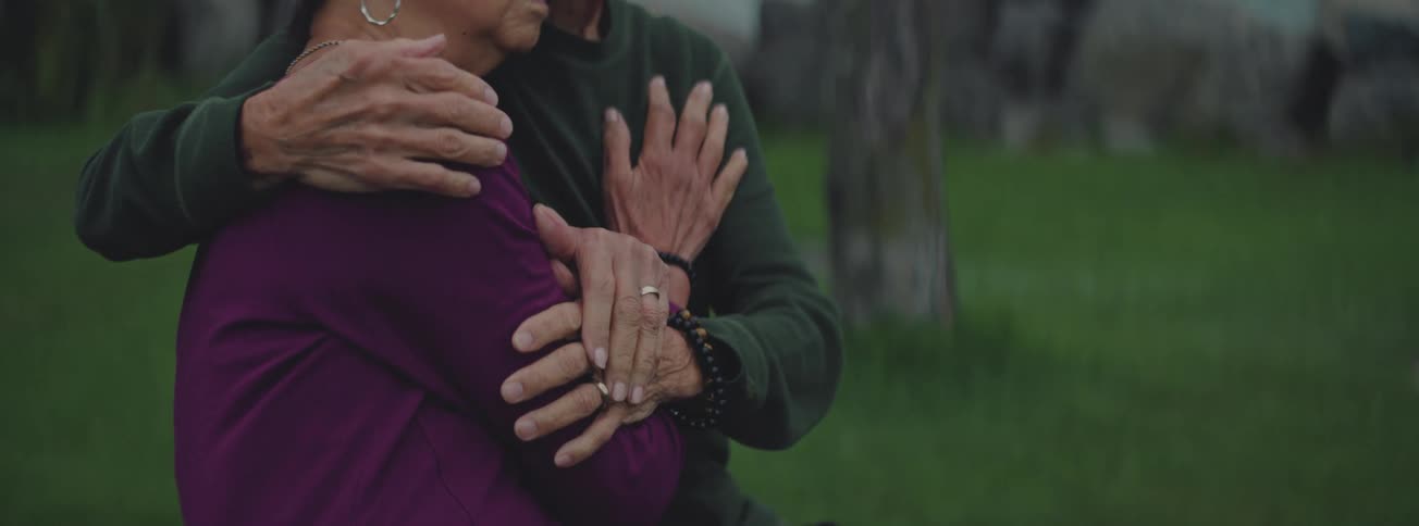 An older couple shares a tender, meditative embrace in a serene outdoor setting, their hands showing a deep connection