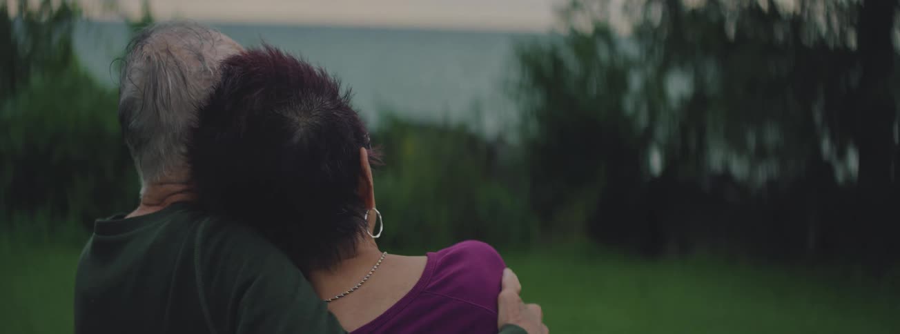 Slow pan of an elderly couple from behind, embracing as they sit together and look out at the water