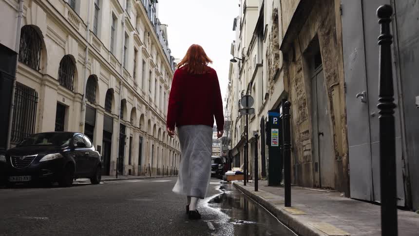A woman with red hair walks in slow motion down a narrow, wet alley in a European city