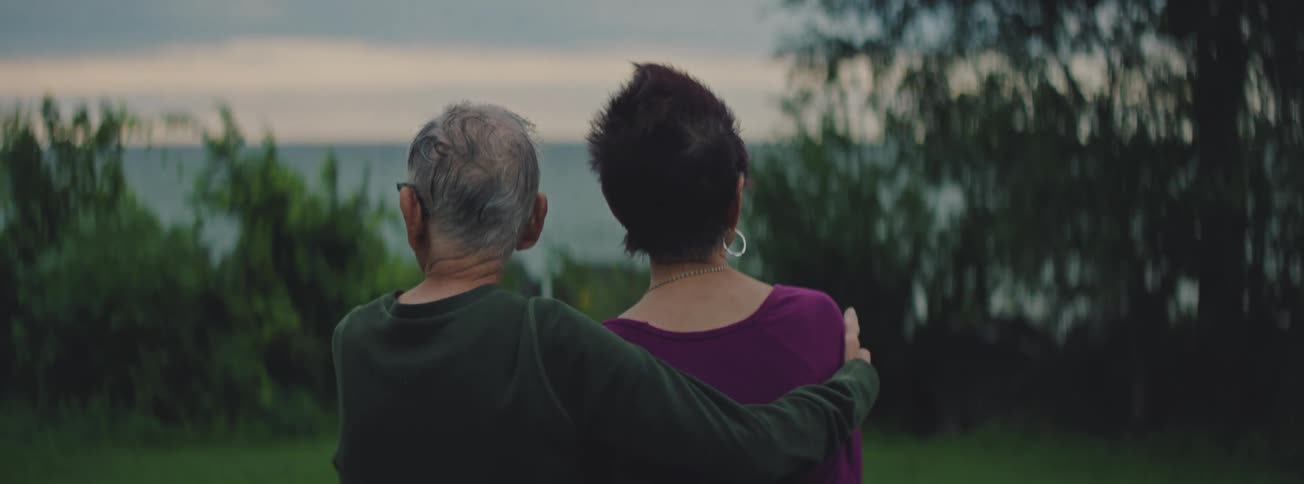 From behind, an elderly couple with their arms around each other talks while looking at a peaceful body of water