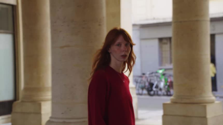 A stylish woman with red hair walks thoughtfully through a classic stone colonnade in a European city in slow motion