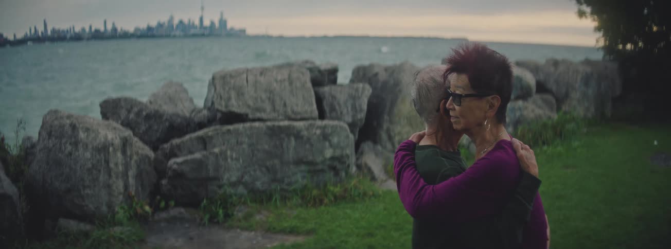 Two senior women share a tender and emotional hug by a rocky lake shore with the city skyline in the distance