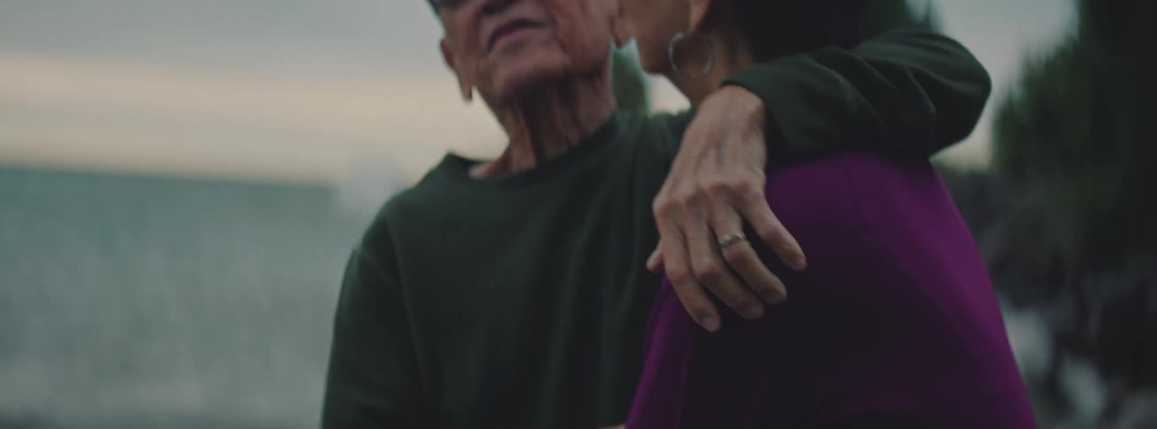A close-up slow-motion shot of an elderly couple sharing a tender embrace and smiling at each other by the water at dusk