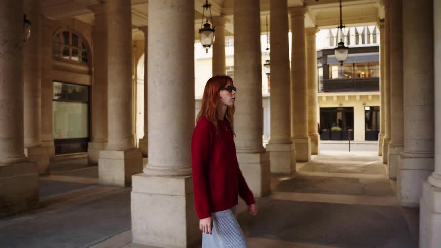 A stylish woman with red hair walks in slow motion through a classic European colonnade