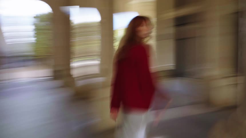 A dreamy, motion-blurred perspective follows a woman in a red sweater as she walks through a grand, sunlit colonnade