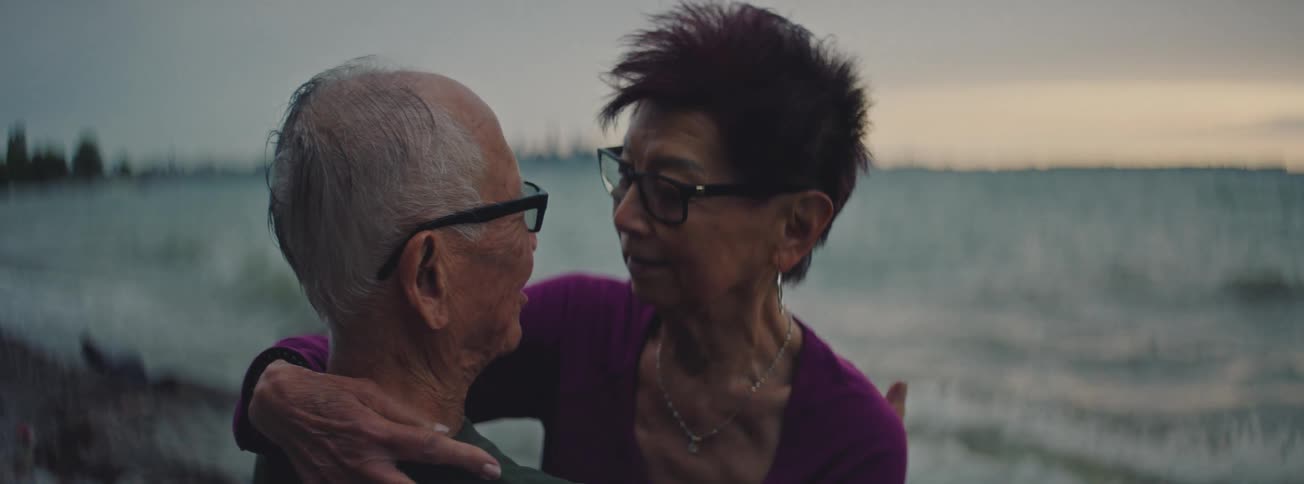 An intimate close-up shot of an elderly Asian couple embracing and talking lovingly by the water's edge at dusk