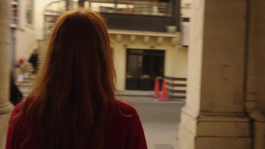 A stylish young woman with red hair and freckles walks thoughtfully through a sunlit European arcade in slow motion