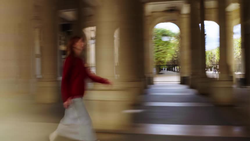 A dreamy motion blur effect captures a woman in a red sweater walking through a sunlit colonnade