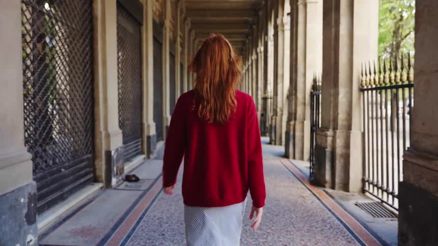 Slow motion shot from behind of a stylish woman with red hair walking down a long, elegant colonnade in a historic city