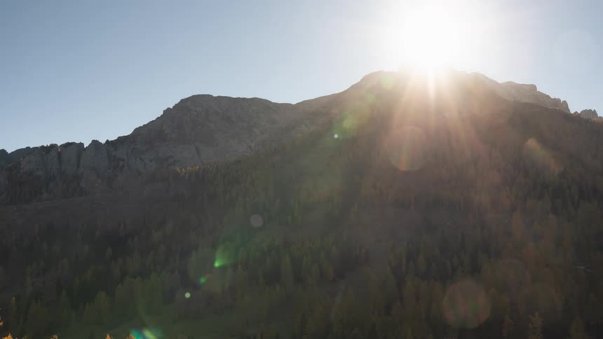 A timelapse of the bright sun shining over a rugged mountain peak and a vast autumn forest below