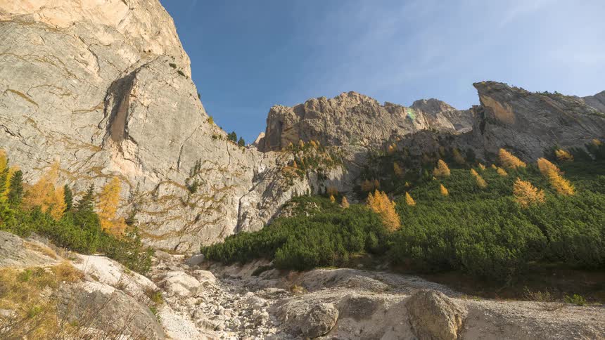 A stunning time-lapse of a rocky alpine valley, with golden autumn larches and green pines under a clear blue sky as shadows move across the landscape