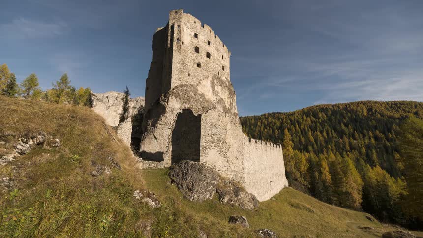 A timelapse captures moving shadows and clouds over the ruins of an ancient stone castle on a grassy hill surrounded by an autumn forest