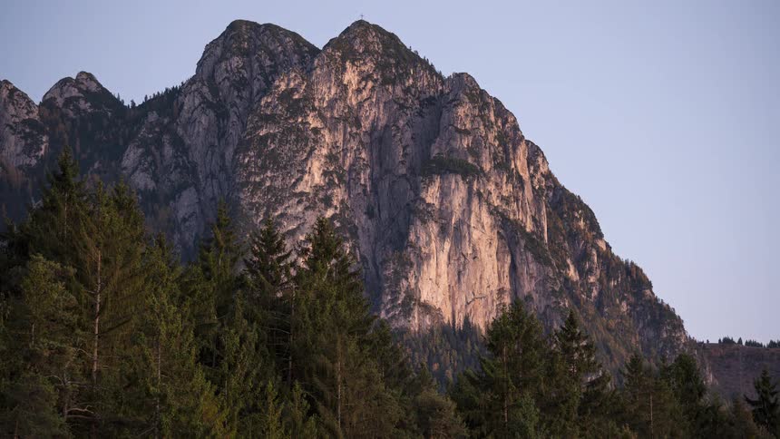 A time-lapse captures the changing light of sunset on a rugged mountain face above a dense evergreen forest