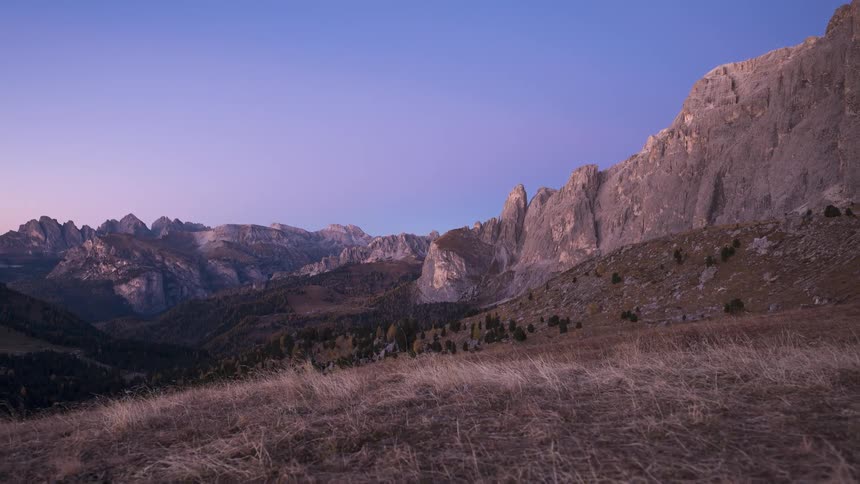 A serene time-lapse captures the first light of dawn washing over a majestic mountain range in the Dolomites