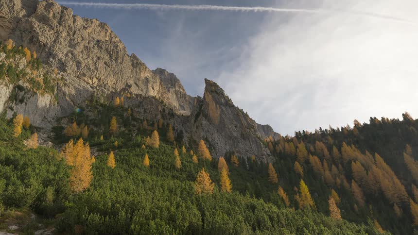 A timelapse of clouds and jet contrails moving across the sky above a rugged mountain peak and a forest of green pines and golden autumn larches