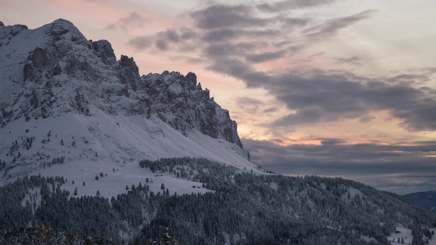 A beautiful time-lapse of a pink and orange winter sunset over a majestic, snow-covered mountain range and pine forest