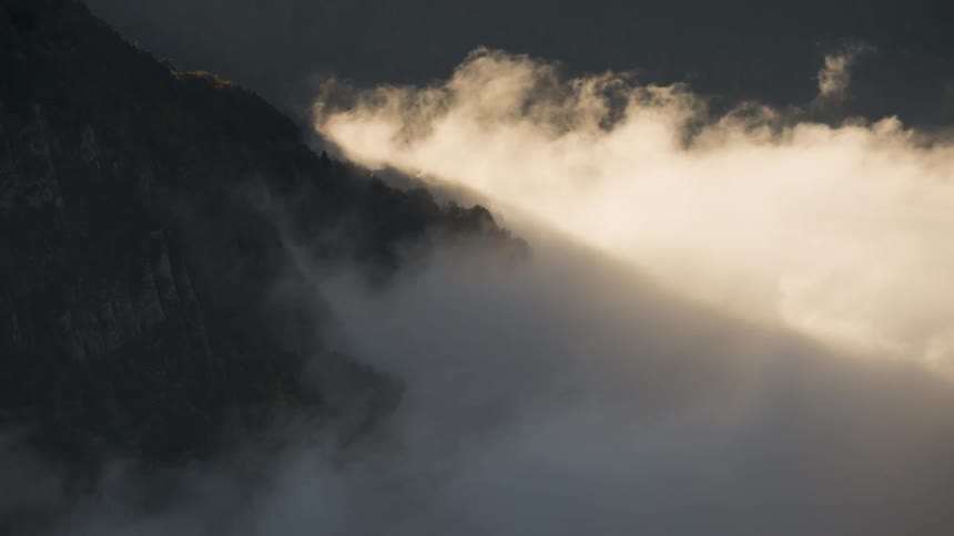 A dramatic timelapse shows a river of thick fog flowing over a dark mountain ridge illuminated by the warm light of sunrise