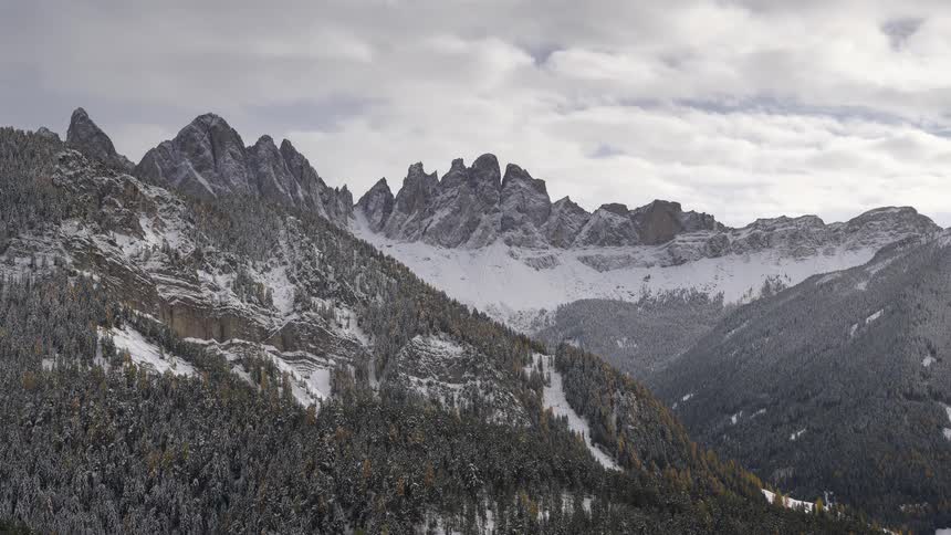 A majestic timelapse captures clouds swirling around the snow-dusted peaks of a rugged mountain range above a dense evergreen forest