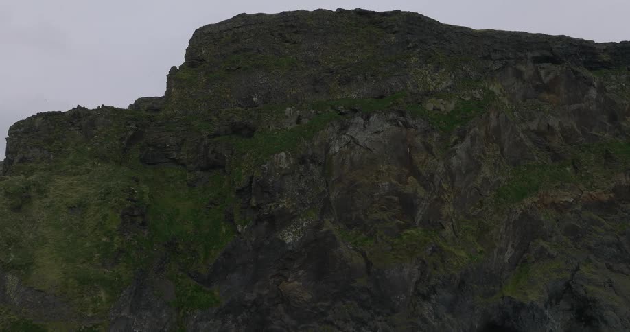 A slow and dramatic aerial shot pans up a rugged, moss-covered cliff on a cloudy day