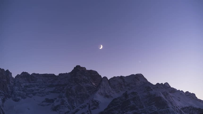 A crescent moon and a bright star hang in the serene twilight sky over a snow-capped mountain range