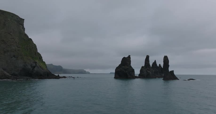 Dramatic basalt sea stacks rise from the dark ocean next to a rugged cliff on a moody, overcast day in Iceland