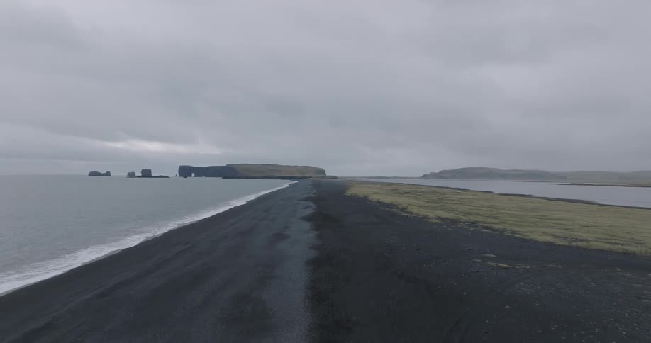 An aerial drone slowly pulls back over a vast black sand beach on the Icelandic coast, revealing the Dyrhólaey peninsula under an overcast sky