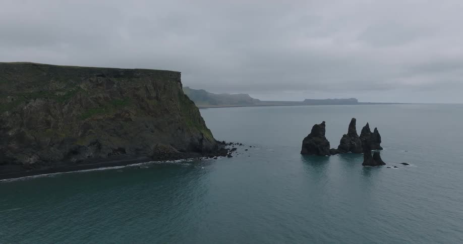 The iconic Reynisdrangar sea stacks stand stoically in the ocean next to the dramatic cliffs of Iceland's southern coast on a cloudy day