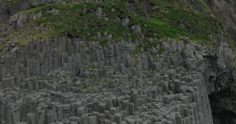 An aerial shot slowly pans across the dramatic, hexagonal basalt column cliffs of Iceland, topped with vibrant green moss
