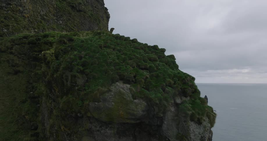 A dramatic aerial shot follows a rugged, green cliff face, revealing the iconic Reynisdrangar sea stacks in the ocean