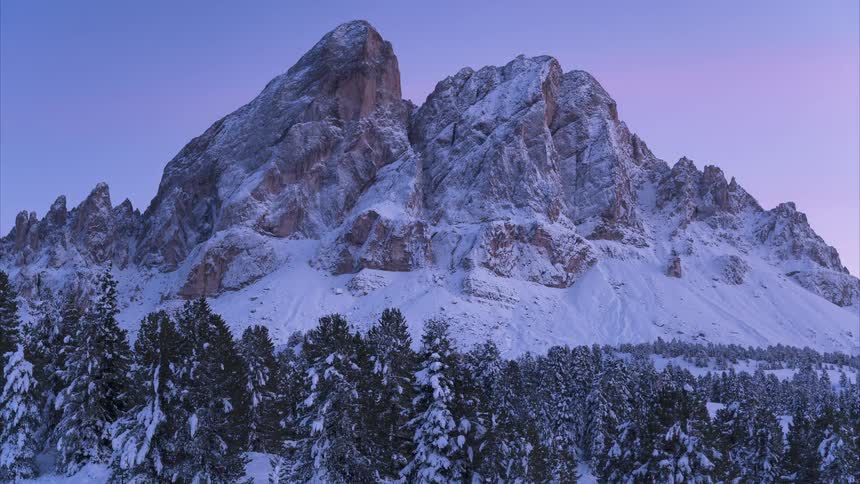 A serene time-lapse captures the changing colors of twilight on a snowy mountain peak above a winter forest