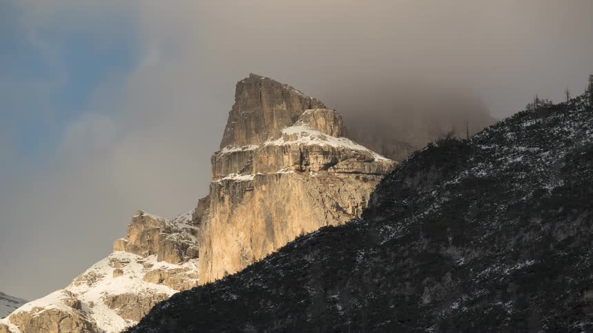 A timelapse captures majestic clouds rolling over a sunlit, snow-covered mountain peak on a dramatic day