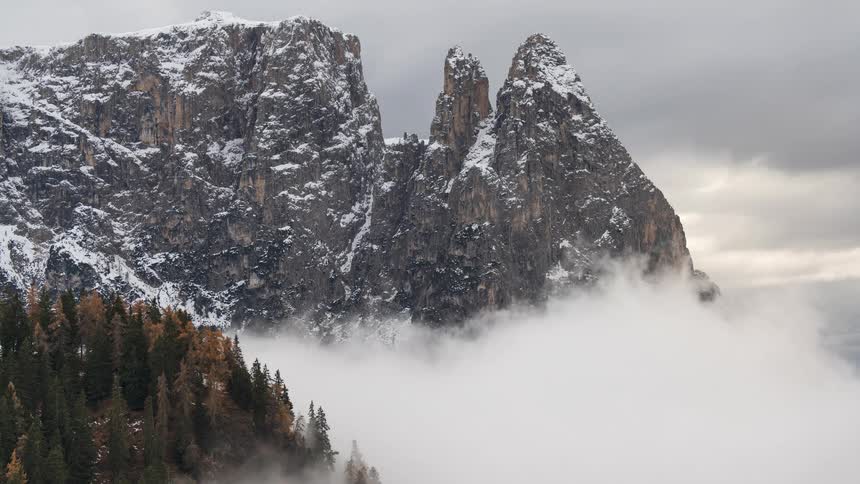 A dramatic time-lapse of thick fog rolling in to cover the base of a majestic snow-capped mountain