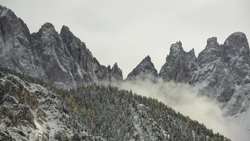 A time-lapse of clouds rolling over majestic snow-covered mountain peaks and a dense pine forest in winter