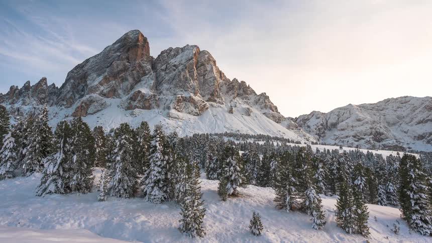 A beautiful time-lapse captures the shifting sunlight and shadows across a majestic snow-covered mountain and evergreen forest