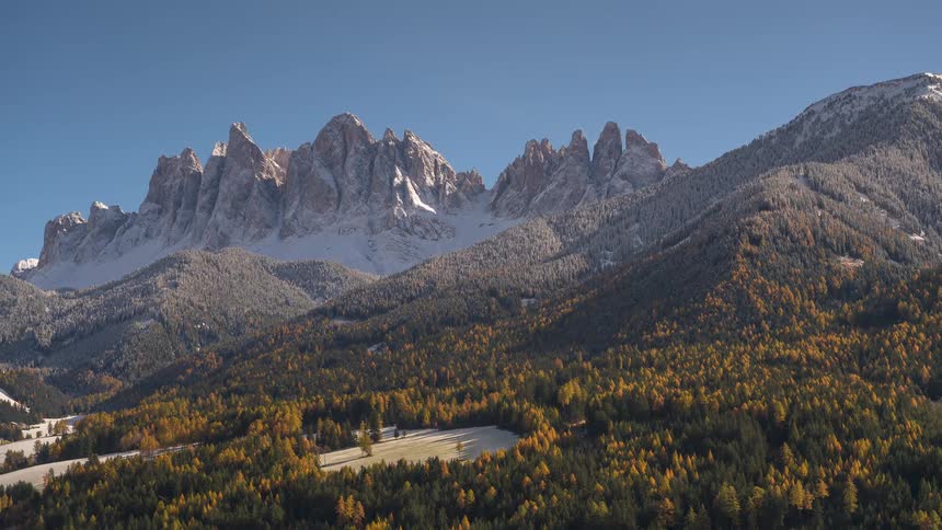 A sweeping aerial shot captures the majestic, snow-dusted peaks of the Dolomites rising above a dense forest of golden autumn larches