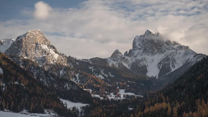 A serene, wide shot shows the majestic, snow-capped peaks of the Dolomites towering over a valley of golden larch and pine trees