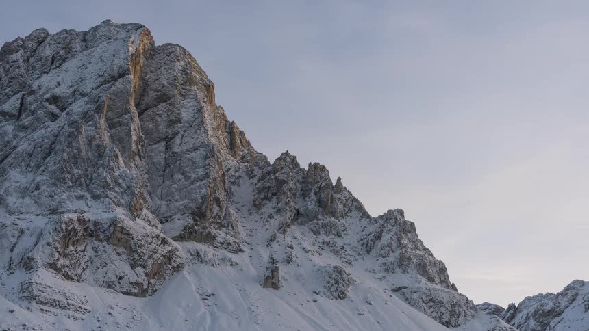 A timelapse captures the last rays of golden sunlight fading from a rugged, snow-covered mountain as evening approaches