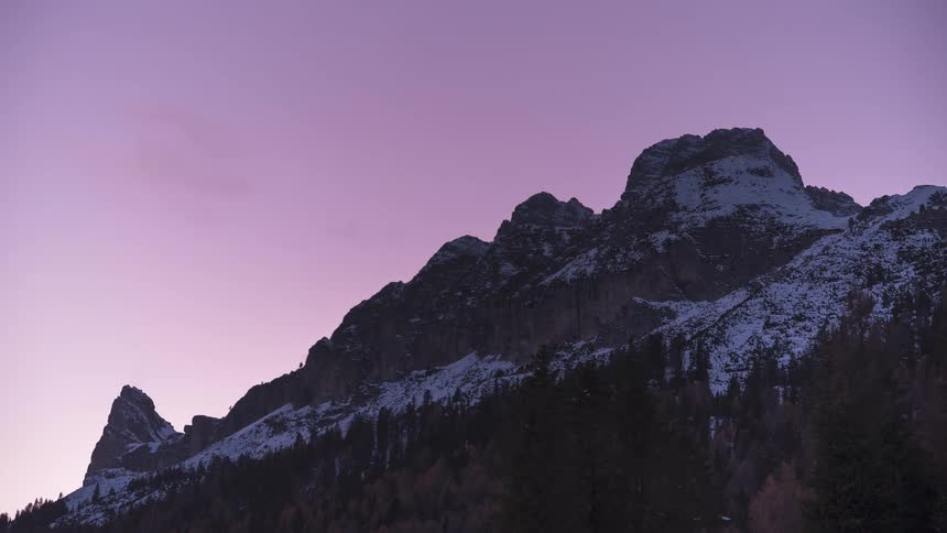 A serene time-lapse shows the twilight sky changing from purple to deep blue behind majestic snow-capped mountains