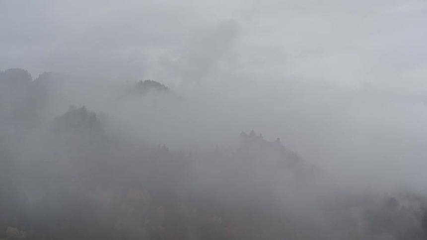 Time-lapse of heavy fog rolling over a mountainous forest, intermittently revealing a mysterious castle and ruins