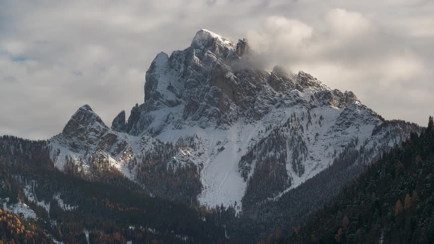 Sunlight illuminates a grand, snow-covered mountain peak as clouds slowly drift across the summit