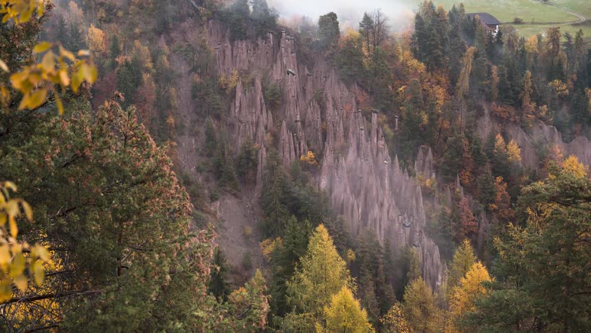 Fog rolls through the valley over the stunning Earth Pyramids of Ritten amidst a vibrant autumn forest in this timelapse