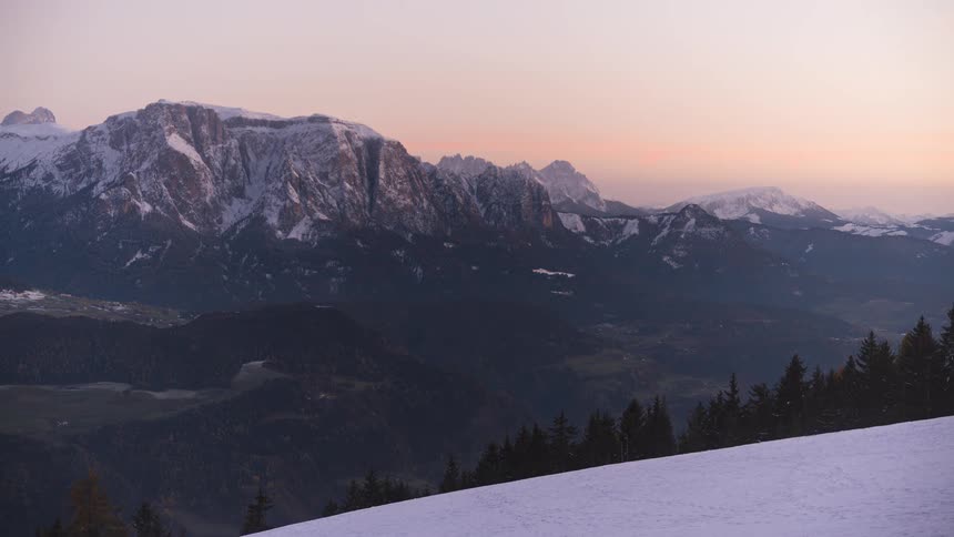A breathtaking day-to-night timelapse captures the sunset casting its final golden glow on a majestic snowy mountain range before fading to reveal the twinkling lights of the valley below