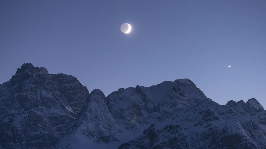A timelapse captures the moon's phases from a sliver of a crescent to a bright full moon over a snowy mountain range at night