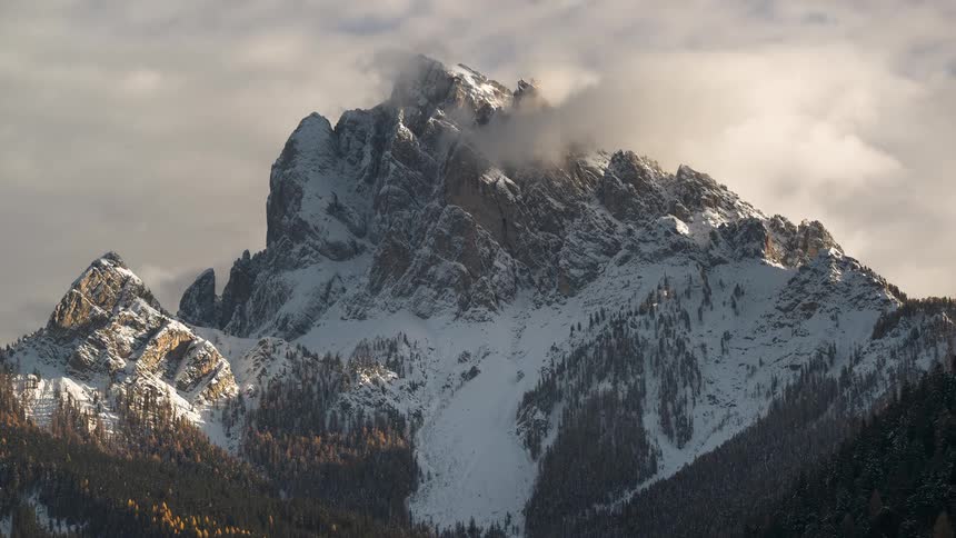 A majestic, snow-covered mountain peak stands tall as clouds drift and swirl around its summit