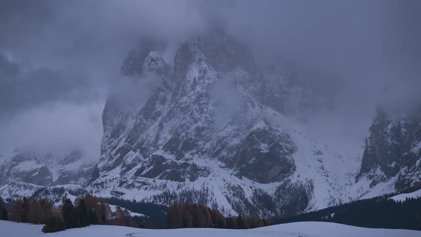 A dramatic time-lapse shows moody clouds swirling around the peak of a massive snow-covered mountain