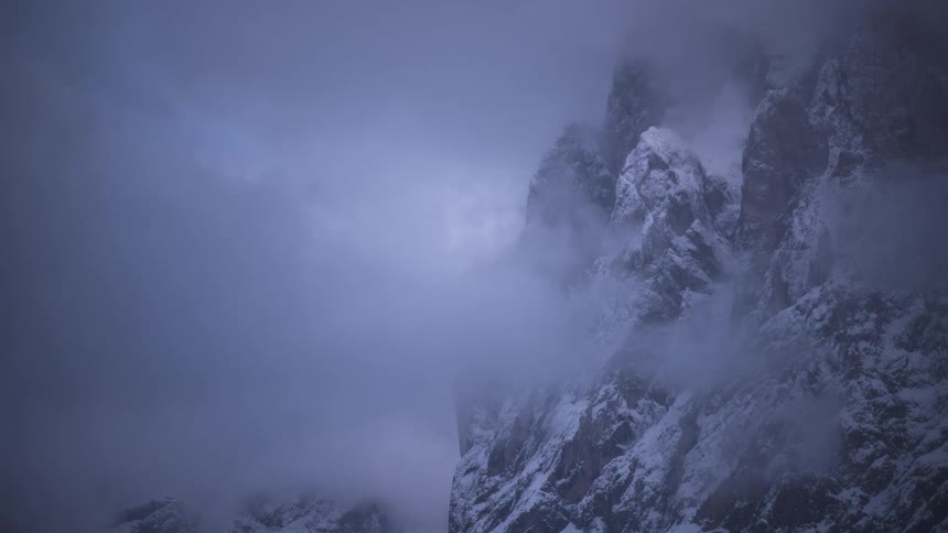 A moody time-lapse shows thick fog and clouds swirling around a dramatic snow-covered mountain peak