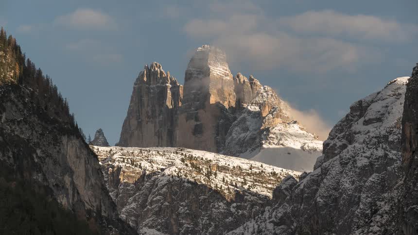 Sunlight illuminates the majestic, snow-covered peaks of the Tre Cime di Lavaredo as clouds drift through the valley