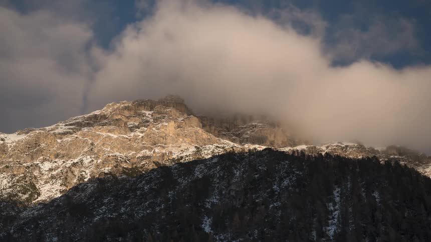 Sunlight illuminates a rugged, snowy mountain peak as clouds drift across the top in a beautiful time-lapse