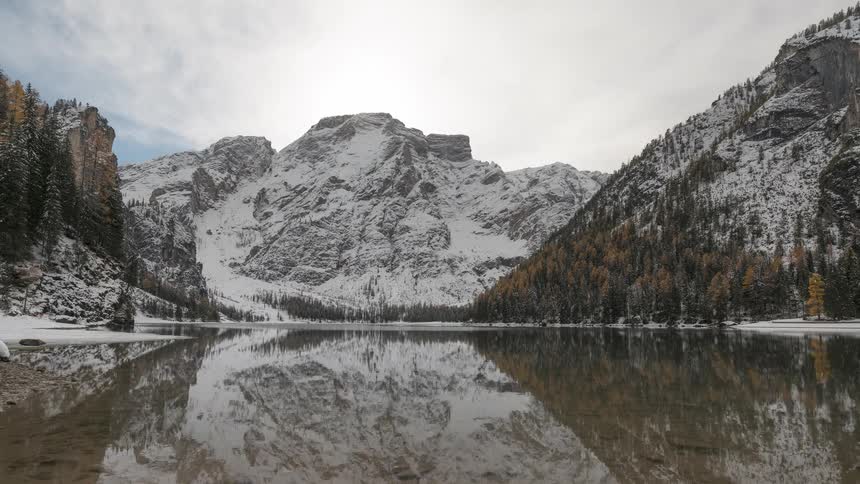 A serene winter landscape shows snow-covered mountains perfectly reflected in the still waters of an alpine lake